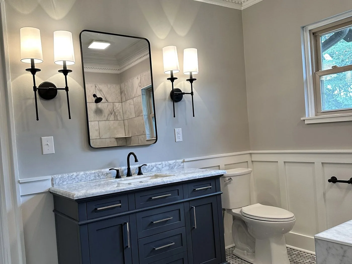 A bathroom with a navy vanity, marble counter, black sconces and a basketweave marble floor