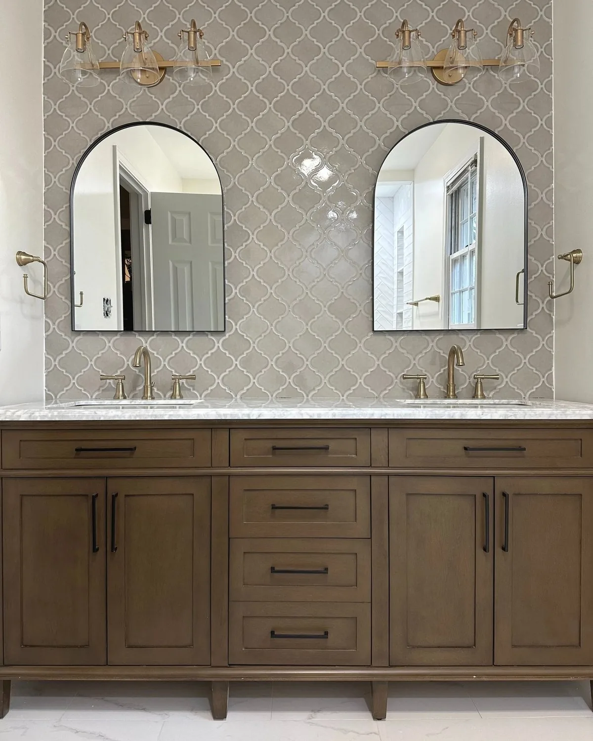 A finished bathroom with arabesque wall tile, two arched mirrors, brass sconces and a wood double vanity