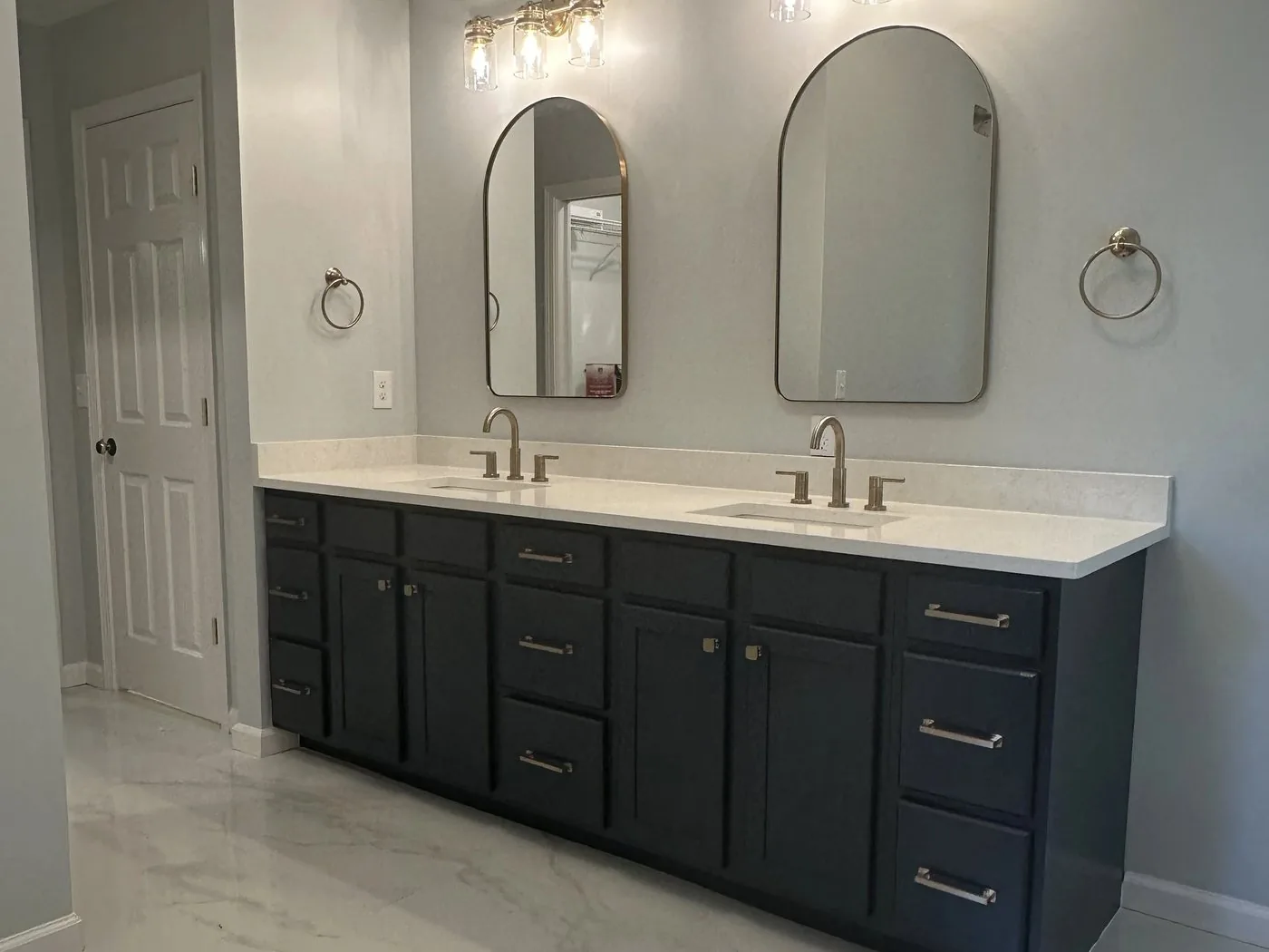 A bathroom with a dark double vanity, arched mirrors, brass sconces and a marble tile floor